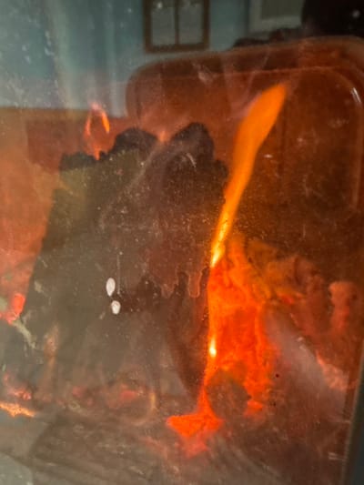 This image presents a close-up view of an active fire contained within an enclosed structure, likely a fireplace or wood-burning stove, observed through a heavily smudged glass panel. The scene is located in Senokos, Bulgaria. Intense orange and yellow flames are prominent on the right side and bottom of the frame, actively consuming dark, solid fuel material, consistent with wood or coal, which is visible in the central and left portions. The glass barrier is significantly obscured by streaks and numerous small, white particulate deposits, indicative of soot and dust accumulation. Reflections on the upper portion of the glass reveal elements of the surrounding indoor environment, specifically a dark rectangular object, possibly a framed picture or window, on a lighter-toned wall, and the curved outline of a reddish-brown object, potentially a piece of furniture. No human subjects are present.