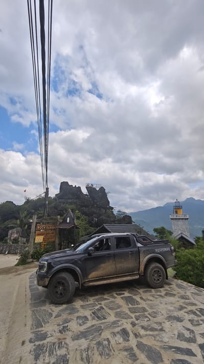 A black, mud-splattered Ford Ranger Raptor pickup truck sits parked on a stone-paved surface. Behind the truck stands a sign that reads "ROCK GARDEN, HOMESTAY, COFFEE, BBQ," written in Vietnamese. A mountainous, rocky landscape looms in the background, with a small lookout platform visible on the peak. A white and yellow lighthouse-like structure is situated to the right of the rock formation. Power lines extend from a pole on the left side of the image. The sky is filled with mostly white and gray clouds. The scene is likely in Tả Van, Vietnam, as indicated by the sign in Vietnamese.