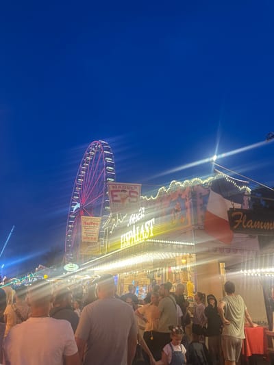 A lively carnival scene unfolds against a dark blue sky. A brightly lit Ferris wheel with red lights spins nearby a busy food stand. The stand has a sign with "Flammkuchen" displayed prominently and a flag. People are milling about, enjoying food and drinks. The crowd appears dense, with many faces and figures partially obscured. The scene is vibrant and full of energy.