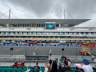 The image shows a view of the Miami International Autodrome, located in Miami Gardens, Florida, United States of America, during the Miami Grand Prix. The track is separated from the seating area by a fence. A large screen displays the event logo and sponsor information. The Hard Rock Stadium is visible in the background. Spectators are seated in the foreground, watching the event. A construction lift with workers is seen on the track level. The environment is overcast with a grey sky.
