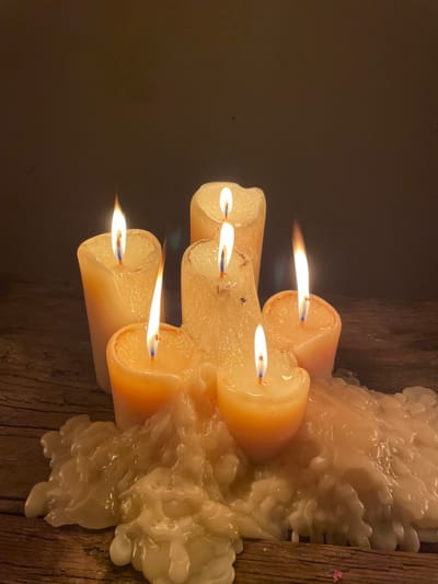 Five lit candles, clustered together, cast a warm glow against a dark background. The candles are cream-colored and round, with flames reaching out from the wicks. Accumulated wax cascades down the side of the candles, solidifying into irregular shapes. The candles rest on a rough wooden surface, its texture visible beneath the wax. The overall scene is dimly lit, with a focus on the flickering flames and the textures of the candles and wood.