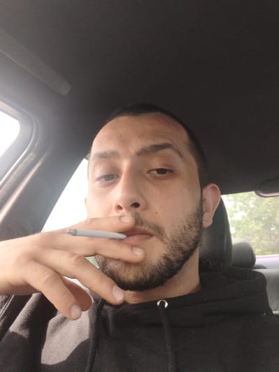 A man is photographed from a low angle, seemingly within a car. He is holding a lit cigarette between his lips and fingers. The car's interior is visible, including the sunroof and a portion of the dashboard. The man is wearing a black hooded sweatshirt. The location, Stafford, United Kingdom, is not evident from the image.