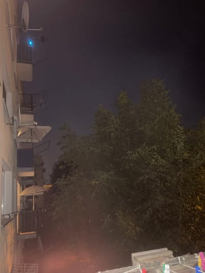 The image depicts an outdoor scene at night, likely from a balcony in Konstantynów Łódzki, Poland. On the left side of the frame, a building facade is visible, featuring several balconies. One balcony has a satellite dish and a small blue light. Another balcony below displays a partially opened umbrella with a striped pattern. Further down, another balcony is visible with laundry hanging on a line.
To the right of the building, a dense cluster of trees with dark green foliage dominates the middle and right sections of the image. The sky above is dark, suggesting nighttime. In the foreground, at the bottom of the image, a section of a rooftop or balcony railing is visible with several clothespins of various colors (pink, green, blue, yellow) clipped to a clothesline. A faint reddish glow emanates from the lower left portion of the image, potentially from an artificial light source or reflections. The overall atmosphere is dark and quiet, characteristic of a residential area at night.
