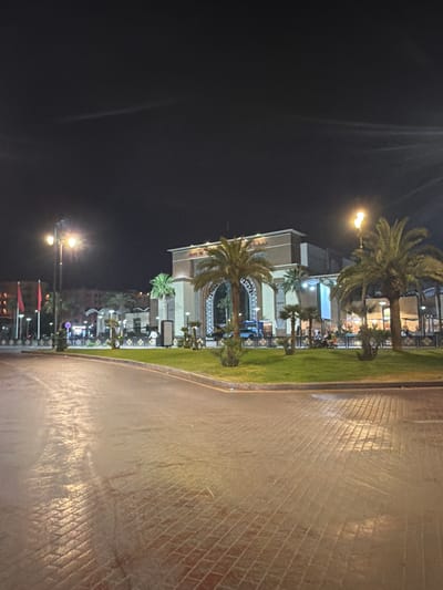 At night in Ouahat Sidi Brahim, Morocco, a brightly lit building with intricate designs and palm trees sits next to a brick walkway and grassy area. Streetlights illuminate the scene, casting light across the architecture and landscaping. People are present on the patio of the building, presumably eating at the restaurant and lounge of the Four Seasons Resort Marrakech.