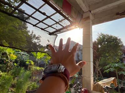 In the city of Karangkebagusan, Indonesia, the image captures an outdoor scene at what appears to be a residential property. A hand, adorned with a watch and bracelets, is raised towards the viewer, with its fingers splayed open. The hand is positioned in the foreground, partially obscuring the view of the environment beyond. Behind the hand, a weathered pergola-like structure with a mesh covering is visible, with sunlight filtering through it, creating a lens flare effect. Beyond the pergola, lush green vegetation, including trees and plants, thrives. In the background, residential buildings with tiled roofs are discernible. The sky is clear and bright, suggesting daylight. The overall impression is one of a sunny day in a tropical or subtropical environment.