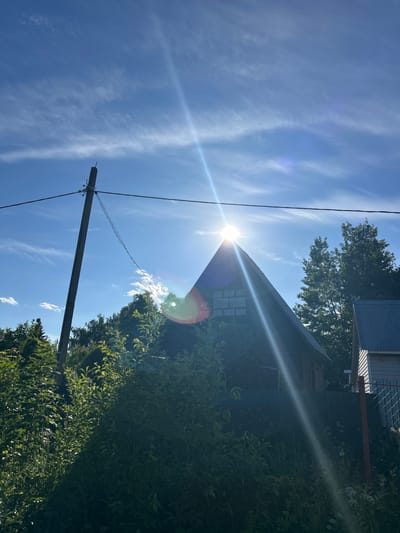 In the city of Moskovsky Literator SNT, Russia, under a bright, sun-drenched sky with wispy clouds, a dark, triangular-roofed house dominates the frame. The sun is positioned directly over the apex of the roof, creating a strong lens flare that bisects the image diagonally from the upper left to the lower right. This flare obscures details of the roof and the windows, which appear as a grid of small panes. Tall green foliage surrounds the house, obscuring much of its base. To the left of the house, a tall, weathered utility pole with wires stretches across the sky. To the right, the corner of another building with a corrugated metal roof is visible, adjacent to a fence. The ground in the foreground is covered in dense, overgrown vegetation and tall grasses. The overall impression is a sunlit, somewhat wild, rural setting.