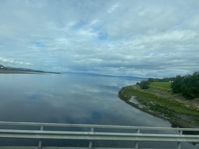 The image depicts a panoramic view of a calm body of water, likely a fjord or large river, under an overcast sky. The water's surface reflects the clouds and the distant, hazy treeline of mountains or hills. In the foreground, a silver metal guardrail runs horizontally across the bottom of the frame, suggesting the vantage point is from a bridge or elevated walkway. To the right, the water meets a muddy, vegetated shoreline with scattered debris. Beyond the shoreline, a lush green field is visible, dotted with trees and two small white houses in the distance. Overhead, several power lines are strung across the sky, creating horizontal lines above the landscape. The overall scene is serene and natural, with hints of human infrastructure. The location is identified as Verdalsøra, Norway.