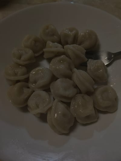 A plate of pelmeni, a Russian dumpling, is shown in a dimly lit indoor setting, possibly in Galonki, Poland. The pelmeni are arranged in a cluster on a white plate, and appear to be cooked and glistening with a light sauce or broth. A fork is partially visible on the right side of the plate, suggesting the meal is about to be eaten or has been partially consumed. The lighting is low, creating shadows and a slightly muted color palette for the food and the plate. The background is out of focus and appears to be a table surface.