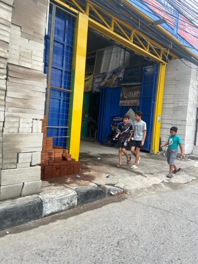 The scene is in front of a building with a yellow and blue striped awning. The building has brick walls with a yellow painted column on the left. There are a stack of red bricks at the base of the column. There are three boys in the scene. Two are walking away from the camera, one is wearing a soccer jersey and shorts and the other is wearing a blue t-shirt and black shorts. The third boy is walking towards the camera and is wearing a green t-shirt and black shorts. The boys are on the sidewalk. There is a motorcycle parked in the background. The scene is located in the city of Bojongsoang, Indonesia, at coordinates -6.972 latitude and 107.636 longitude.