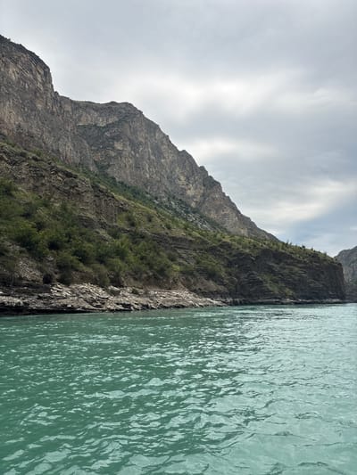 A wide-angle, eye-level shot captures a rugged mountain landscape alongside a vibrant, turquoise river. The jagged, rocky mountains dominate the left and central portions of the frame, with steep, weathered slopes that show layers of rock and sparse vegetation. The river flows in the foreground, its surface textured with ripples and reflections, extending towards the right side of the image where the mountains continue, narrowing the view. The sky above is overcast with muted, grey clouds, casting a soft light over the scene. The image appears to be taken from a boat or a low vantage point on the river, looking towards the mountains and the riverbank. The overall impression is one of natural grandeur and a remote, untouched environment, possibly a river canyon. The location is identified as Zubutli, Russia, suggesting a natural geographical feature within or near this area. There are no people or visible man-made structures in the photograph.