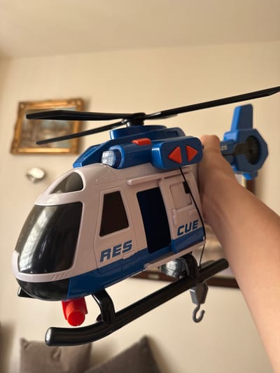 The image displays a close-up, angled shot of a person's hand holding a toy rescue helicopter indoors. A fair-skinned right hand is seen grasping the upper part of the helicopter.
The toy helicopter is predominantly white and blue, with black main rotor blades and a black tail boom. A small grey hook is attached to the end of the tail boom. Red cylindrical components are visible on the sides of the fuselage, and two orange arrow-shaped buttons are set into a blue panel on the top surface of the helicopter. The words "CUE" are printed in blue capital letters on the top, and "RES" is printed in blue capital letters on the side of the white fuselage.
The background is a blurred, light-colored (possibly beige or off-white) wall, suggesting an indoor setting. Faint, out-of-focus details include a gold-framed picture and a small, round, light-colored object, implying a domestic environment. The activity captured is a hand holding the toy, possibly for play or display. The specific geographical location of Oldham, United Kingdom, is not visually discernible from the content of the image itself.
