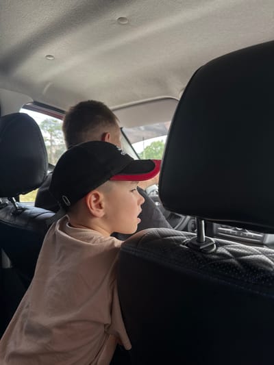 A medium shot shows a young boy in a black baseball cap with red trim and a light pink t-shirt sitting in the back seat of a car, looking to his right. His face is partially obscured by his cap and the car's headrest. Behind him, the back of a man's head and shoulders are visible. The interior of the car is dark, with black leather seats and a light-colored headliner. Through the window, trees and foliage are visible. The scene is set in Berezovaya Staritsa, Russia.