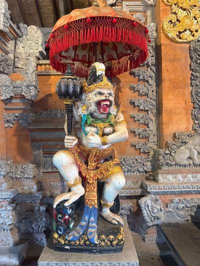 A multi-colored statue of a fierce, humanoid figure with a monkey-like head and bared fangs stands prominently within a traditional Balinese architectural setting in Batuan, Indonesia. The figure, possibly a deity or guardian, is depicted in a dynamic, squatting pose, clutching a large, dark, club-like mace in its right hand. It features a light cream-colored body, adorned with a golden crown, a green chest adornment, and intricate gold and red patterned loincloths and leg wraps. Its lower legs are painted blue with gold accents, resting on a black and gold base.

Above the statue is a ceremonial umbrella (payung) with a decorated, reddish-orange canopy and numerous dangling red tassels. The immediate environment consists of intricately carved grey stone architectural elements, including pillars, cornices, and panels featuring swirling motifs and stylized animal figures, such as a crouching lion-like creature visible on the right. The walls behind these carvings are constructed from reddish-orange brick or textured plaster. Wooden ceiling beams are visible at the top. The scene is illuminated by natural light, casting distinct shadows.