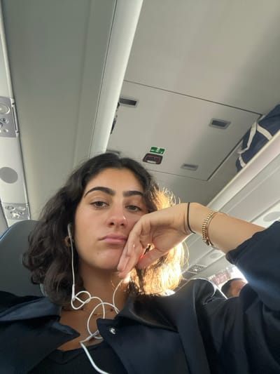 The image depicts the interior of a passenger transport vehicle, likely an airplane or a long-distance bus/train, characterized by overhead compartments, recessed lighting, and an emergency exit pictogram on the ceiling. The specific location within the city of Pombal, Portugal, cannot be determined from the visual information presented in the image.
A young adult female is prominently featured in the foreground, seated and looking directly into the camera. She possesses dark, wavy, shoulder-length hair and an olive complexion, with a neutral to slightly pensive facial expression, her mouth slightly downturned. She wears white wired earbuds, gold-colored hoop earrings, and a delicate gold-colored necklace with small red elements. Her attire includes a dark top and a dark collared shirt or jacket worn over it. Her right hand, adorned with a black elastic hair tie and a gold-colored chain-link bracelet, supports her right cheek/chin.
The background shows the vehicle's interior: a light-colored ceiling with overhead panels and compartments. A blue and white striped bag is stowed in an overhead compartment on the right. Natural light streams into the vehicle from the right side, illuminating the subject's right side. A partial view of another passenger's head is visible in the far right background. A dark grey seat back is visible to the subject's left.