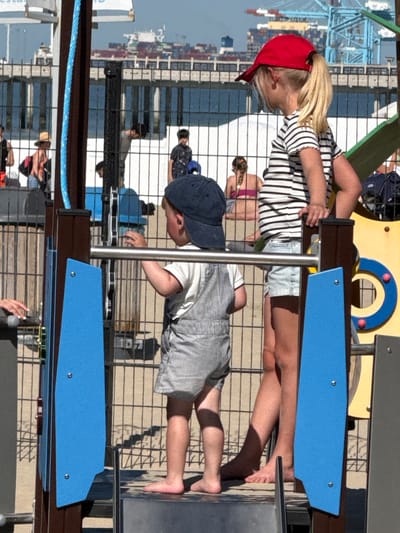 Here is a detailed description of the image:
Two children are playing on a playground structure. One child, wearing a red baseball cap and a striped shirt with jean shorts, is standing. The second child is wearing a blue baseball cap, a white shirt, and striped shorts. The children are on a playground with blue and brown elements. In the background is a cityscape with cranes and shipping infrastructure, and a harbor on the coast of Blankenberge, Belgium. Fencing is present behind them, separating the play area from the harbor. There are other people in the background, some on the playground structure.