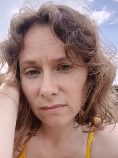 The image shows a close-up, selfie-style shot of a woman with light brown, wavy hair. She has a fair complexion with freckles and is looking slightly off-camera with a neutral expression. Her left arm is visible resting near her face. She is wearing a yellow top. The background appears to be outdoors with a cloudy sky. There is no discernible urban environment or specific location information provided to indicate a connection to Revda, Russia.