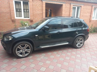 A dark green BMW X5 SUV is parked on a patterned paved driveway in front of a brick building. The building has white-framed windows and a wooden door. The SUV's windows are tinted, and the car has a roof rack. The paving stones are a mix of pink and light gray. The scene appears to be in a residential area, likely in Zyazikov-Yurt, Russia, given the context. There are no people visible in the image.