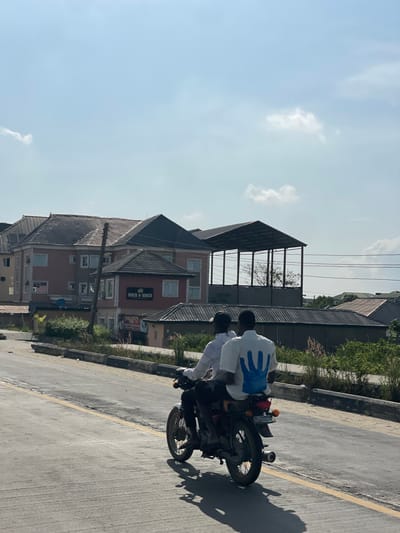 The scene is set on an asphalt road under a bright, clear sky with minimal cloud cover in Igando Oloja, Nigeria. Two adult males are observed from behind, riding a dark-colored motorcycle away from the viewer. The driver wears a light-colored, long-sleeved shirt and a dark cap. The passenger wears a white short-sleeved shirt featuring a prominent blue handprint graphic on the back. The motorcycle has a red taillight and a license plate partially visible on its rear fender. The road surface is gray asphalt with a faded yellow line demarcating lanes, and a low concrete curb or barrier runs along the right side.

In the mid-ground and background, a row of buildings and structures is visible. To the left, a multi-story building with a light reddish-brown facade and a dark gray tiled roof stands. In front of it, a smaller, two-story structure with a dark tiled roof displays a sign that reads "HACK & WIACK" on its dark-colored lower facade. Further to the right in the background, an open-sided structure with a dark roof and metal framework is present. Overhead utility lines traverse the scene horizontally. Sparse vegetation, appearing as dry grasses and shrubs, occupies the ground between the road and the background structures. Other residential-style buildings with dark roofs are partially visible further in the distance.