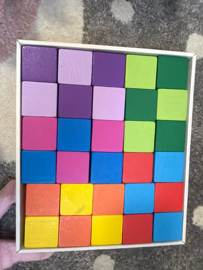 The image shows a top-down view of a white box filled with colorful wooden blocks. The blocks are arranged in a grid pattern of 5 rows and 5 columns. The colors of the blocks, from top to bottom and left to right, are: purple, light purple, pink, blue, orange, yellow, and red. The box is placed on a gray and white striped carpet. There is no visible indication of the location being Krasnyi Krym, Russia.