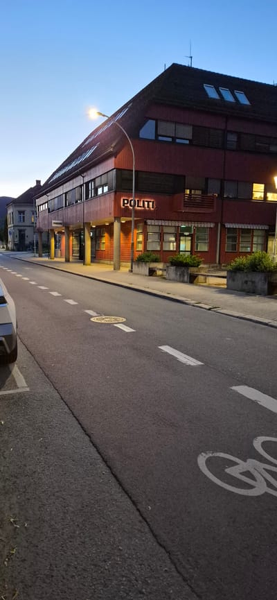 The image shows a police station in Lillehammer, Norway, with a street in the foreground. The building is red with a dark roof and large windows. The word "POLITI" is illuminated on the front of the building. The street is paved and has white lane markings. There is a bicycle lane marking visible on the right side of the street. The sky is clear and blue, indicating it is either early morning or late evening. A street lamp is visible on the left side of the building. No people are visible in the image.