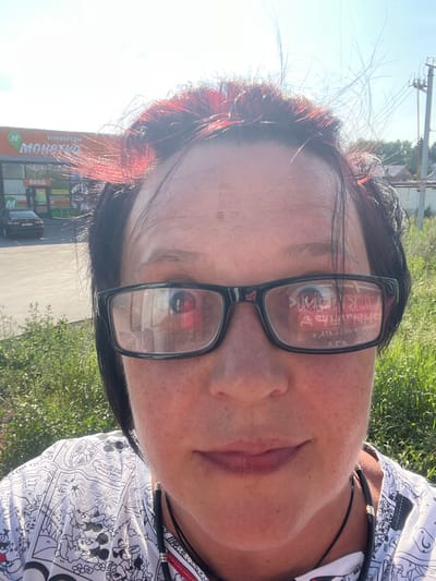 The media file is a close-up, sideways selfie of a person wearing glasses. The subject has dark hair with reddish highlights, and is wearing a white t-shirt with a black and white cartoon pattern, possibly featuring Mickey Mouse. A black cord is visible around the person's neck. The background consists of green grass and a bright sky, suggesting an outdoor setting during daylight hours. Reflections in the glasses show text in Cyrillic, which appears to be from a sign in the background. Visible text includes "УНИВЕРСАМ" (supermarket) and "ВХОД" (entrance). The overall impression is of a casual, sunny day. Based on the provided location context, the scene is likely in Yarkovo, Russia.