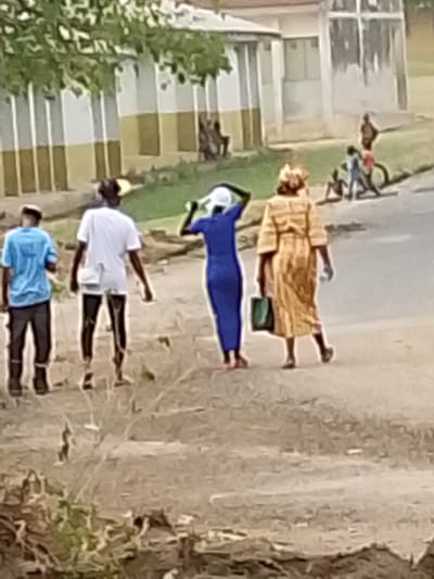 The image depicts an outdoor daytime scene, likely on an unpaved road or path, with several individuals and buildings in the background. In the immediate foreground, four people are walking away from the camera.

On the far left, a person wearing a light blue short-sleeved shirt and dark pants is visible. Next to them, a second person is wearing a white short-sleeved shirt, dark pants or shorts, and a light-colored bag slung across their body. The third individual from the left is a woman dressed in a form-fitting, royal blue long-sleeved garment, possibly a dress or a top and pants. She is wearing a white hat or head covering, and her hands are raised towards her head, suggesting she might be adjusting the hat or shielding her face from the sun. The fourth person on the right is a woman wearing a long, flowing, light golden or tan-colored dress or robe with a matching headwrap. She carries a dark green bag in her left hand.

The ground beneath them appears to be a dry, earthen path. In the mid-ground on the right side of the image, a group of several smaller figures, likely children, are visible. Some are standing, others are seated or crouching, and one is near a bicycle, suggesting they are playing or interacting.

In the background, there is a building characterized by vertical stripes or panels, alternating between white and a muted yellow or light brown color. Some green foliage and trees are visible in the upper left portion of the image and scattered around the scene. The lighting indicates it is daytime. Due to the image's blurriness, specific facial features or fine details are not discernible. No text is visible in the image.