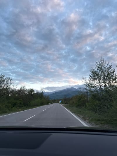 The image shows a view from inside a vehicle, likely a car, looking out onto a road in a mountainous area. The road has a single white line marking the lane. Trees line both sides of the road, and in the distance, there are visible mountains partially obscured by clouds. The sky is filled with clouds, and the lighting suggests it is either dawn or dusk. The location is possibly near Mezdreya, Bulgaria, based on the environmental characteristics.