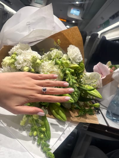 A photograph shows a hand with an engagement ring resting atop a bouquet of white and green flowers. The bouquet is wrapped in brown paper and white tissue. The hand has light skin with short, manicured nails. The ring features a dark, square gemstone surrounded by smaller, possibly diamond, stones in a silver setting. The background suggests the interior of a vehicle, likely an airplane or train, due to the presence of a window with a view of the outside. Additionally, there is a water bottle visible in the background. The location is likely not in the city of Domodossola, Italy, based on the environmental cues in the background.