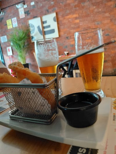 Two glasses of beer sit in a metal rack. The top glass is full, while the bottom one has a smaller pour. A black condiment cup is next to the top beer. Fried food fills the metal rack beneath the beer glasses. A white, plastic serving station supports the rack. A brick wall appears in the background, along with table decor.