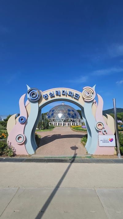 The image depicts the entrance to a park in Cheokbeonjeong-ri, South Korea. The entrance features a large, colorful archway with abstract wavy designs in pastel colors. The Korean text "농심테마파크" (Nongshim Theme Park) is displayed in blue lettering across the top of the arch. Through the archway, a geodesic dome structure with blue and clear panels is visible, situated within a landscaped area. The ground in front of the dome is a paved path. To the right of the archway, a sign reads "THEME PARK BIG HEART GEOJE." The environment is outdoors with a clear blue sky and some foliage and flowers visible. A shadow of a lamppost is cast on the ground in the foreground.