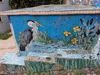A mosaic-covered structure stands outdoors in Soure, Portugal. The dominant feature of the artwork is a depiction of a gray heron, rendered in fragmented tiles, set against a blue background. Additional details include stylized yellow flowers and green vegetation, also composed of mosaic pieces. The structure has multiple tiers, each adorned with mosaic designs that visually represent water and foliage. The overall impression is a decorative, outdoor installation, likely a public art piece.