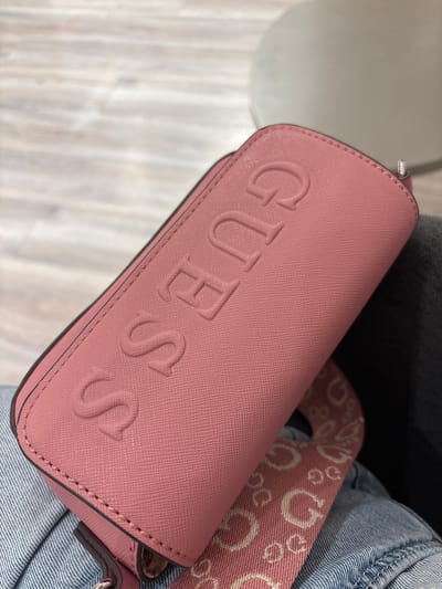The image shows a close-up of a pink Guess brand purse. The word "GUESS" is embossed on the front of the purse. The purse has a strap with the Guess logo printed on it. The purse is resting on a pair of blue jeans. The setting appears to be indoors. The image lacks specific environmental cues indicating it is in Rionegro, Colombia.