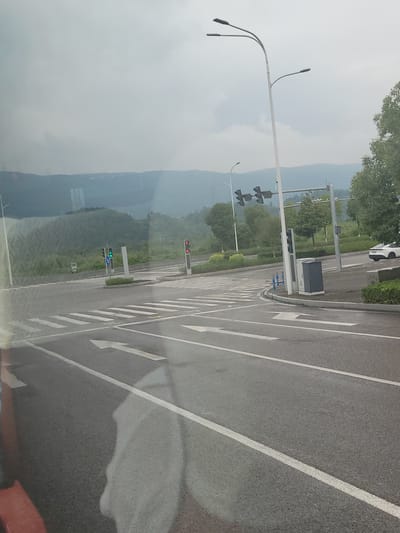 The image shows a road with traffic lights and street lamps. The road is asphalt and has white lane markings. There are several traffic lights visible, some showing red and some showing green. A pedestrian crossing is marked with white stripes. In the background, there are trees and mountains, suggesting a natural landscape. The sky is overcast, indicating a cloudy day. The time of day appears to be daytime. The setting looks like a suburban or rural area with a road junction. No people are clearly visible, but a white car is partially seen on the right side of the road. There is no visible text in the image. The overall scene depicts a normal day at a road intersection with traffic control systems in place.