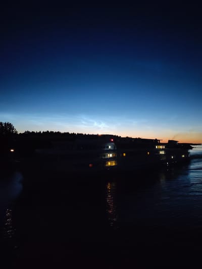 The image depicts a dark, atmospheric scene of a body of water with a vessel at dusk. The sky transitions from a deep blue at the top to lighter hues near the horizon, suggesting the twilight hours. A long, dark silhouette of what appears to be a large boat or ferry runs horizontally across the middle ground. Several illuminated windows are visible on the vessel, casting a warm glow onto the water. To the left of the vessel, a cluster of trees is silhouetted against the sky. The water in the foreground is dark and appears rippled, with reflections of lights from the vessel. The overall impression is one of a quiet, serene evening by the water. The location is identified as Sud'bitsy, Russia.