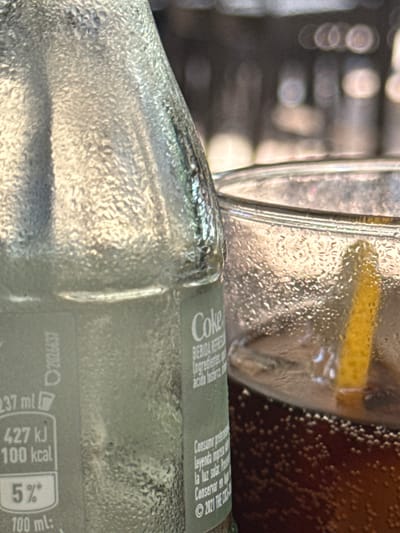 This image captures a close-up of condensation-covered glass bottles and a glass, suggesting a cold beverage is being served or has recently been consumed. The focus is on the textures and reflections of the water droplets on the glass surfaces.

The subjects are primarily objects: a glass bottle, a drinking glass, and likely a bottle of soda. The bottle in the foreground has a label with visible text including "Coke," suggesting it's a Coca-Cola product. Other legible text on the label includes nutritional information like "427 kJ 100 kcal," and a date "2021 THE COCA-COLA COMPANY." The scene is likely indoors or in a shaded outdoor area given the absence of direct sunlight and the cool, refreshing appearance. The lighting appears to be soft, highlighting the condensation. The overall mood is one of refreshment and possibly a casual social setting. The cropped nature of the image prevents a clear view of the surrounding environment, but the condensation implies a cool drink in what could be a warm climate.