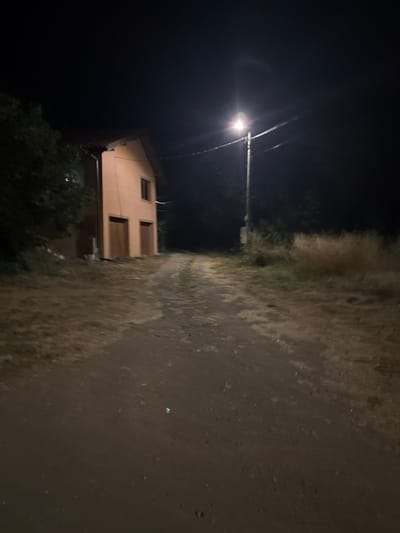 The image depicts a rural scene at night in Kableshkovo, Bulgaria. A dirt road leads away from the viewer towards a two-story house with a light-colored facade and a dark roof. The house has two large, garage-like doors on the ground floor and a single window on the upper floor. To the left of the house, dark foliage is visible. On the right side of the road, tall, dry grass and shrubs are illuminated by a nearby streetlamp mounted on a pole. The sky is dark, indicating nighttime. The overall impression is one of a quiet, isolated location under artificial light.