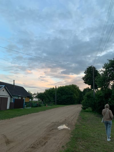 In the village of Bleychitsy, Belarus, at dusk, a woman with blonde hair pulled back in a bun walks away from the camera on the right side of a dirt road. She wears a brown jacket, light blue jeans, and white sneakers, with a light-colored crossbody bag. The road stretches into the distance, showing tire tracks and a small puddle reflecting the sky. On the left side of the road, a house with a white brick facade and a dark roof has a brown garage door. Adjacent to it is a smaller building with green and yellow siding. A few trees are visible behind the house, and a larger cluster of dense green trees forms a wall on the right side of the road, extending into the background. A utility pole with multiple wires stands prominently amidst the trees. The sky above is cloudy, with hues of pale pink and orange from the setting sun peeking through the grey clouds.