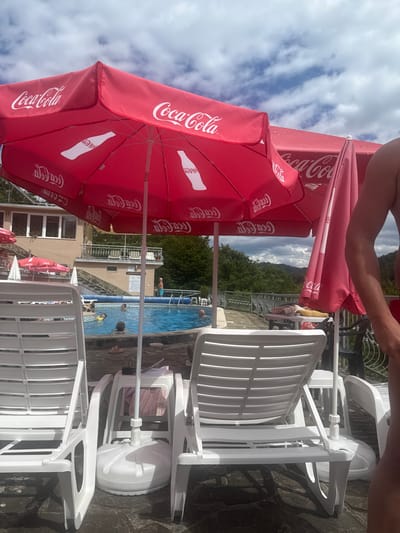 This is a medium-shot, eye-level, vertical, outdoor photograph taken on a cloudy day at the Shipkovo Mineral Baths in Shipkovo, Bulgaria. The foreground features two white plastic lounge chairs with one partially visible on the right edge of the frame, occupied by a person whose torso and arm are visible. The chairs are set on a dark, patterned stone patio. In the middle ground, a bright blue swimming pool is visible, with several people swimming and one person standing at the edge. Two large red Coca-Cola branded umbrellas are prominently displayed, one open and angled towards the viewer, the other partially closed and behind it. Several other red and pink umbrellas are visible in the background. Behind the pool and umbrellas is a multi-story building with balconies and windows, its facade is a light tan color. The background is filled with lush green trees and rolling hills, with a cloudy sky overhead. The overall scene depicts a leisure area at a mineral bath facility.