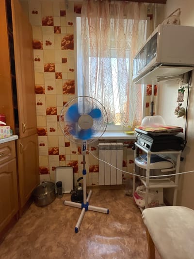 The image depicts an indoor scene, likely a kitchen, with a brown wooden cabinet on the left and a window with sheer curtains on the right. A standing fan with blue blades is positioned in the center of the room, and to its right, a white shelving unit holds various items including kitchen appliances and containers. A radiator is visible against the wall beneath the window. The flooring is a patterned linoleum, and the walls are decorated with a tile pattern featuring cherries. A large pot, a kettle, and a white rectangular tray are on the floor near the cabinet. A chair with a light-colored cushion is partially visible in the foreground on the right. The lighting suggests it is daytime, with light entering through the window. The overall impression is a lived-in domestic space.