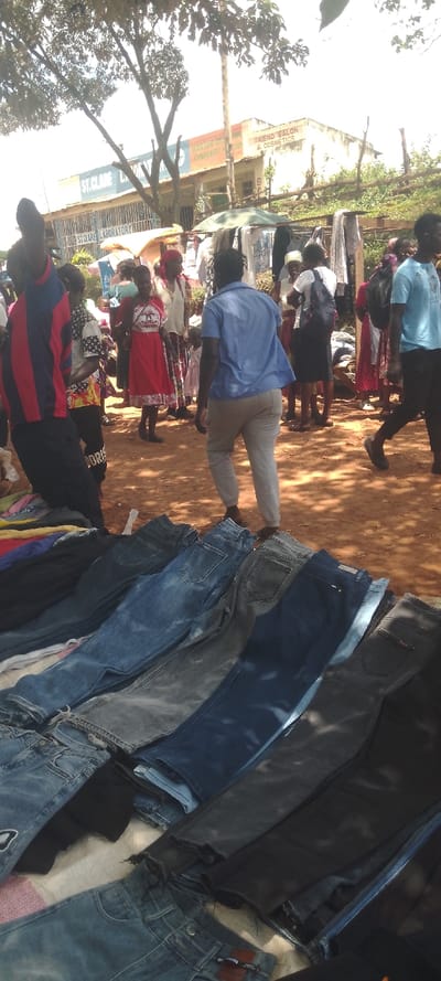 The image depicts a bustling outdoor market scene in Chemuche ward, Kenya. In the immediate foreground, multiple pairs of denim jeans in varying shades of blue and black are laid flat on a surface, likely a vendor's stall. The ground throughout the visible area is composed of reddish-brown dirt.
Approximately fifteen to twenty individuals are present across the mid-ground and background, predominantly adults. A person wearing a light blue short-sleeved button-up shirt and light-colored pants stands centrally in the mid-ground, facing away from the camera. To their left, another individual is partially visible wearing a red and blue striped shirt. Further back, a person in a red dress with white patterns stands, facing right. On the far right, a person in a light blue t-shirt and dark pants walks leftward. Other individuals are scattered, engaged in standing, walking, and browsing activities amidst market stalls.
In the background, market stalls display various clothing items on racks and under rudimentary shelters. Behind these stalls, a row of commercial buildings is visible. One building prominently features "ST. CLAIRE LABORATORY" signage. An adjacent building displays signs for "MARTAM COLLEGE" (partially visible, likely "MARTAM COLLEGE OF EMBROIDERY HOME") and "FRIEND SALON & COSMETICS." A rough wooden fence runs along the background behind the buildings and market. Overhead, large trees with green foliage cast dappled shadows across the scene under bright daylight conditions.