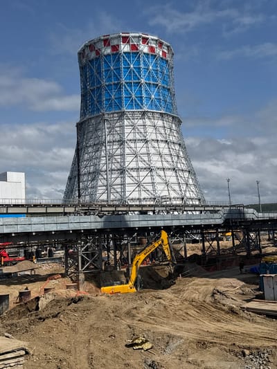 The image depicts a construction site in Chulman, Russia, dominated by a large, partially constructed cooling tower. The tower has a metallic lattice structure, with the upper portion adorned with a pattern of blue and red panels. Below the tower, a complex network of scaffolding and elevated walkways is visible. The foreground is a muddy, excavated area where an excavator, marked with the "SHANTUI" logo, is actively working. Other construction equipment, including a red crane and a yellow forklift, are also present. Several workers, dressed in construction attire, can be seen throughout the site, some operating machinery and others supervising. The environment is characterized by a partly cloudy sky and a generally industrial setting.