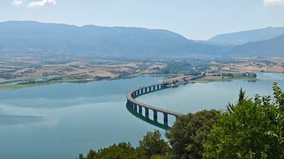 A wide-angle, high-angle shot captures a scenic landscape dominated by a large body of turquoise water, likely a reservoir, with a long, curved bridge spanning across it. The bridge, supported by numerous pillars, carries a road with several vehicles visible. The water reflects the sky and the bridge, creating a symmetrical effect.
In the background, rolling hills and mountains form a hazy backdrop, with scattered agricultural fields in various shades of green and brown. A town or village is visible nestled among the fields and hills in the distance. The sky is a pale blue with a few scattered white clouds.
In the foreground, lush green trees and foliage frame the bottom of the image, adding depth and a natural element to the scene. The overall impression is one of a serene and vast natural landscape complemented by human infrastructure. This location is the Servia-Rapsani Bridge over the artificial lake of Polyfytos, located in the Servia Municipal Unit, Kozani Regional Unit, Greece.