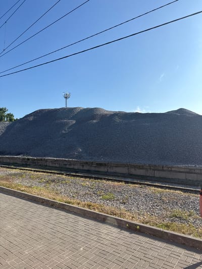 The image depicts a large pile of crushed stone situated next to a railway track. A cell tower is visible on top of the pile. The scene is outdoors, under a clear blue sky. In the foreground, a paved walkway is present, with a concrete curb separating it from the railway line and the crushed stone. Power lines stretch across the upper portion of the image. No people are clearly visible in the frame. The location is identified as Arzamas, Russia.