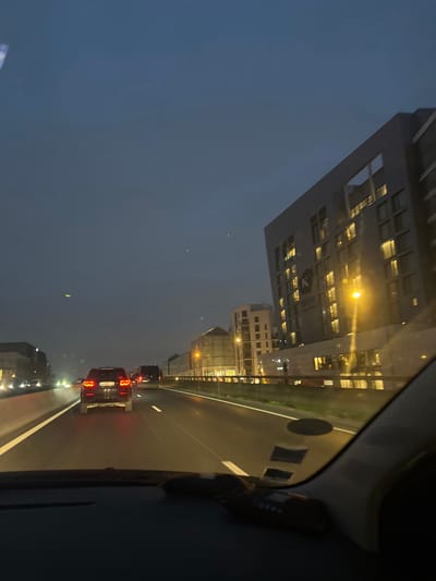 The image captures a view from inside a vehicle at dusk or night. Buildings are visible to the left, illuminated with lights against a dark sky. The road stretches ahead, marked by lane lines and dotted with traffic, indicated by the red taillights of vehicles. The right side of the image shows the car's interior.