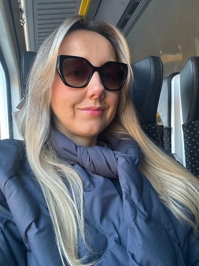 A woman with long blonde hair is sitting in a train car. She is wearing sunglasses and a dark blue jacket. The seats in the train are black leather. The train car appears to be relatively empty. There is no verifiable information to indicate the location in Kalngale, Latvia.