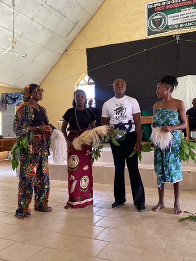 This media file depicts an indoor scene with several individuals, likely at an event in Dutsen Kato, Nigeria. The lighting suggests it could be daytime, with natural light possibly coming from a window or open space.
The foreground features two women, one wearing a vibrantly patterned, traditional outfit and the other in a black top and a dark, patterned skirt. Both are holding leafy branches and appear to be part of a procession or performance. The woman in the patterned outfit is also wearing sunglasses. Behind them, another woman is visible, dressed in a white t-shirt with text and dark leggings, holding similar leafy branches. Further back, a woman in a blue and green patterned wrap is standing.
In the background, a banner is visible with text that includes "FELLOWSHIP OF CHRISTIAN", "NEXT GENERATION", "MOTTO: CARING FOR THE TOTAL SPIRIT, SOUL AND BODY", "VOM BRANCH", and "1 Thess. 5:23". This suggests the event is likely a religious gathering or a community event organized by a Christian fellowship. The overall atmosphere seems celebratory or ceremonial, indicated by the attire and the props. The floor is tiled, and the walls appear to be a light color, possibly painted. The presence of the banner and the traditional attire points towards a cultural and religious context within Nigeria.