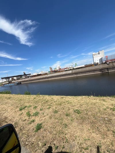 This image depicts an industrial waterfront scene on a clear, sunny day in Neuss, Germany. The perspective is from a slightly elevated position, looking out over a calm body of water that appears to be a river or canal. On the far bank of the water, there is a row of industrial buildings and facilities, including warehouses, silos, and various cranes. Some of the buildings are white with red accents, and one prominently displays the text "ADAMS" in dark lettering.
The waterfront itself is a concrete embankment lined with what looks like railway tracks and various pieces of industrial equipment. There are also some shipping containers visible along the dock. A large crane, likely used for loading and unloading cargo, is a significant feature of the scene.
The foreground of the image is dominated by a dry, grassy embankment. The grass is a light brown and yellow color, indicating it might be summer or a dry season. A shadow is cast on the grass, suggesting the sun is high in the sky, implying it is midday. In the bottom right corner, a portion of a car's side mirror and window are visible, hinting that the photo was taken from a vehicle.
The sky is a vibrant, clear blue with a few wispy white clouds scattered across it. The overall impression is one of an active industrial area on a bright, pleasant day. There are no people visible in the frame, and the focus is on the industrial landscape and the natural elements of the sky and grass.