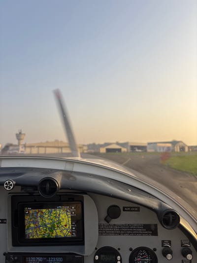 An interior view from the cockpit of a small aircraft captures a scene on a taxiway or runway at Toussus-le-Noble Airport (LFPN), Toussus-le-Noble, France. The ambient light suggests either sunrise or sunset, characterized by a warm, hazy glow near the horizon.

In the foreground, the aircraft's gray dashboard is prominent. A mounted tablet-like device displays a detailed topographical navigation map, showing the time as 17:53. The screen indicates menu options such as "Zoom Out," "Zoom In," and "Direct To." To the right of the screen, a power outlet is labeled "ALIM. 12V DC," and a placard details aircraft operational specifications, including approval for VFR day/night operations, prohibition of flight in known icing conditions, and restrictions on aerobatic maneuvers. Below the main dashboard, a Garmin radio panel displays "120.750" as the active communication frequency and "123.500" as standby. Other instruments are partially visible, including one labeled "ALT FLORA" and a circular engine instrument with red markings.

Through the windshield, the aircraft's propeller is visible in the midground, blurred, indicating the engine is operating. A paved taxiway or runway extends into the distance, bordered by green grass. A small pink marker post is present on the right side of the taxiway.

The background features airport infrastructure, including a light-colored control tower with a cylindrical base and observation deck positioned to the left. Multiple hangars and other airport buildings, generally light-colored, are visible under a sky that transitions from light blue at the zenith to a warm, hazy yellow-orange near the horizon. No individuals are visible.