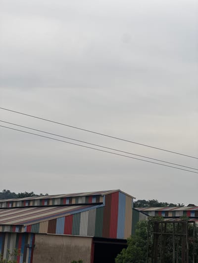 This image captures an outdoor scene featuring buildings with distinctive multi-colored facades under an overcast sky. The original image is rotated 90 degrees clockwise, so describing it in its upright orientation:
On the right side of the frame, two large structures, likely buildings or warehouses, stand out with their exteriors covered in corrugated metal panels. These panels are painted in a striking array of horizontal stripes featuring colors such as red, blue, green, brown, and various shades of grey and white. One building is prominent in the foreground, with another similar structure visible slightly behind it.
Behind and to the right of the buildings, dense, green foliage and trees are abundant, some appearing to be palm trees, suggesting a tropical or subtropical climate. A dark, angular metal structure, possibly a fence or an architectural element, is partially visible through the trees on the far right.
Stretching diagonally across the upper left portion of the sky are three thin, dark power or utility lines. The sky itself is a uniform, light grey, indicating an overcast day or atmospheric haze. There are no people or discernible activities taking place, presenting a static and quiet scene. No text is visible within the image. The setting is indicated to be Ozubulu, Nigeria.