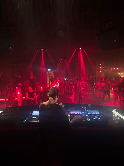 The image depicts a dimly lit nightclub scene in Ban Bang Thao, Thailand, dominated by vibrant red lighting. In the foreground, a DJ stands with their back to the camera, operating a DJ console with illuminated screens and various controls. Their hands are visible on the equipment. Behind the DJ, the nightclub floor is filled with a crowd of people dancing and socializing, illuminated by beams of red light cutting through the haze. Multiple speakers are mounted on the ceiling. The atmosphere appears energetic and lively, characteristic of a nightclub setting.